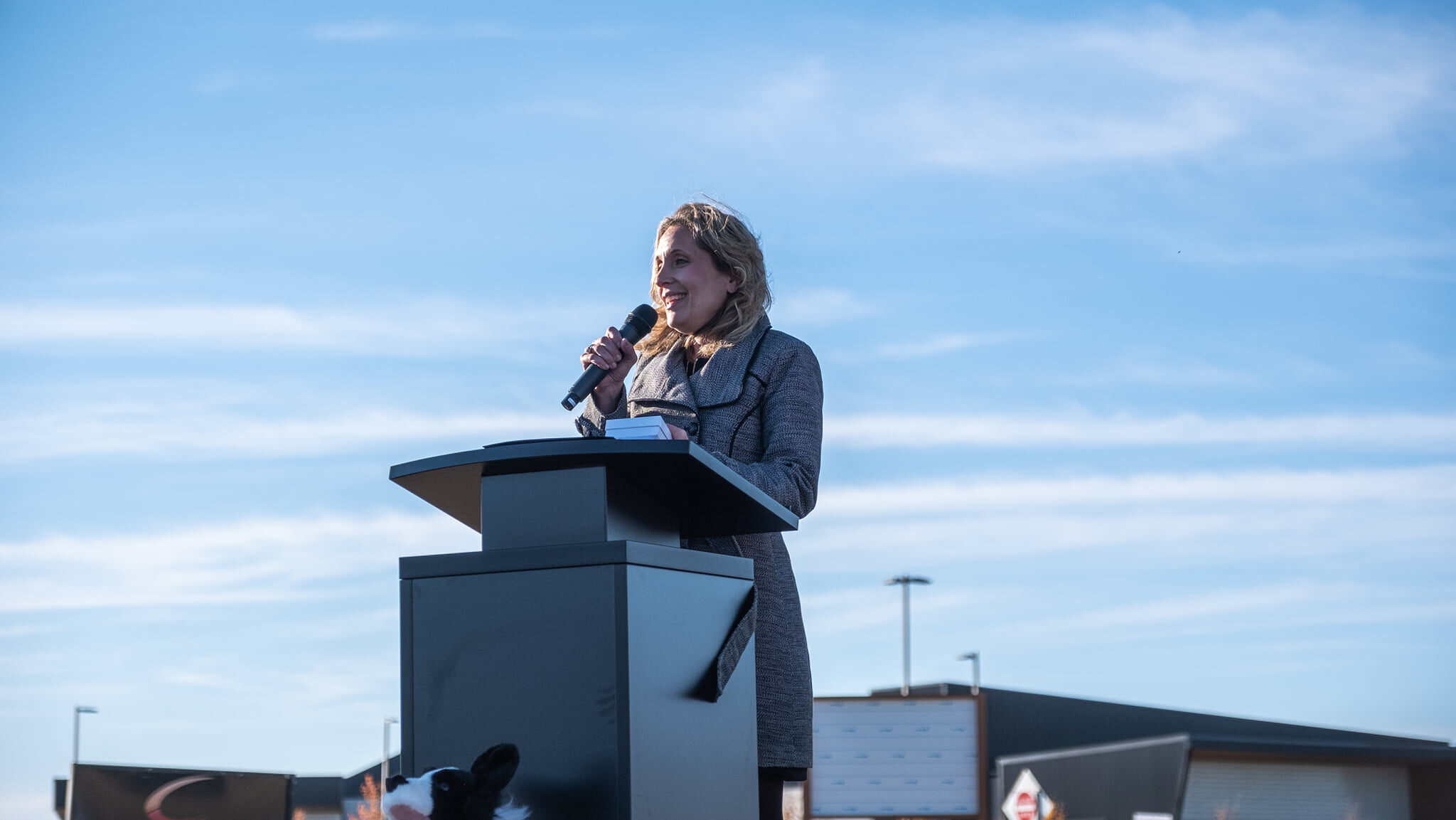 A woman speaks at an outdoor podium on a sunny day. A dog stands beside the podium. There is a building and a sign in the background.