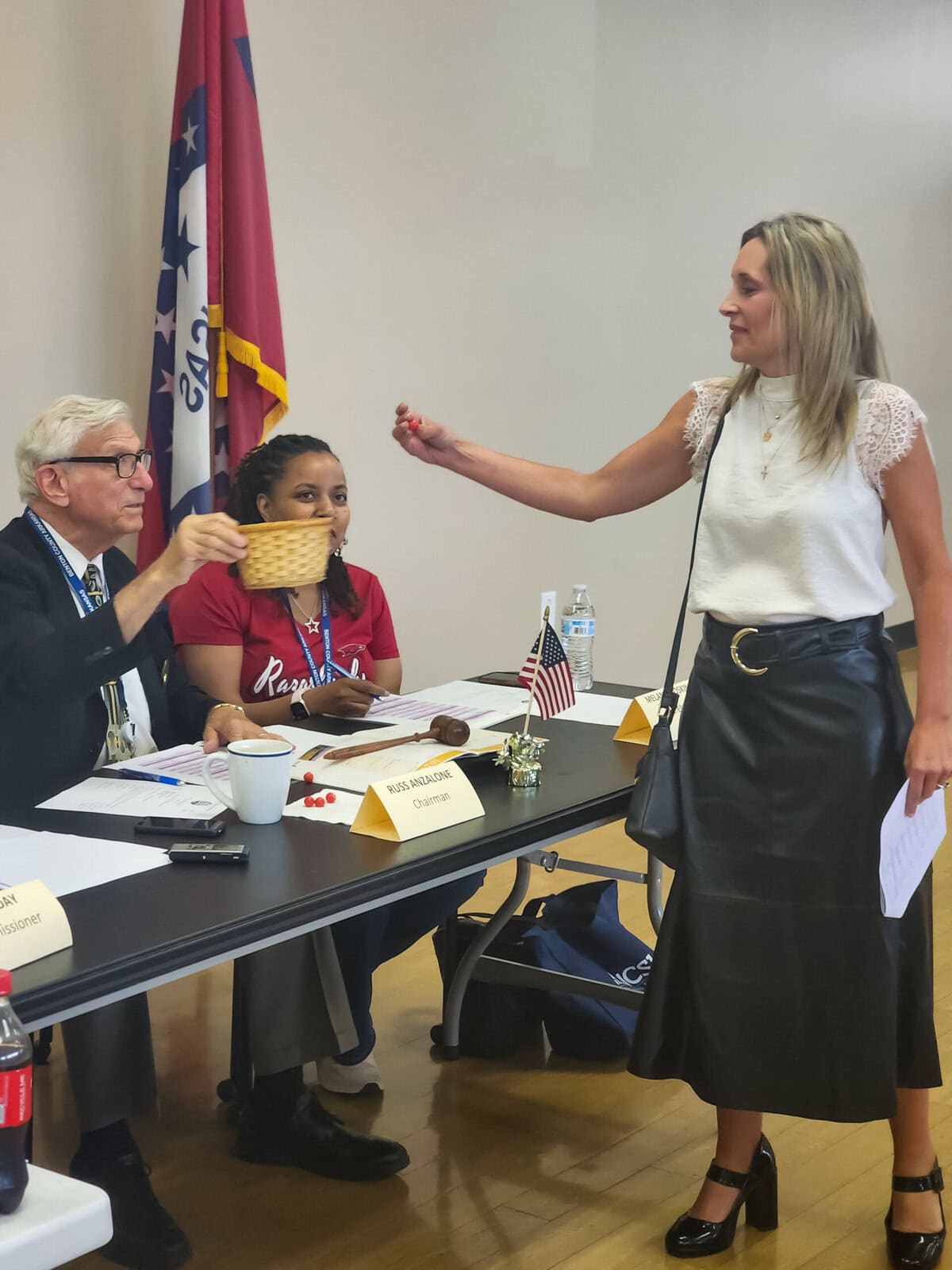 Mayor Orman stands in front of a table with three seated individuals, holding a piece of paper. The table has flags, papers.