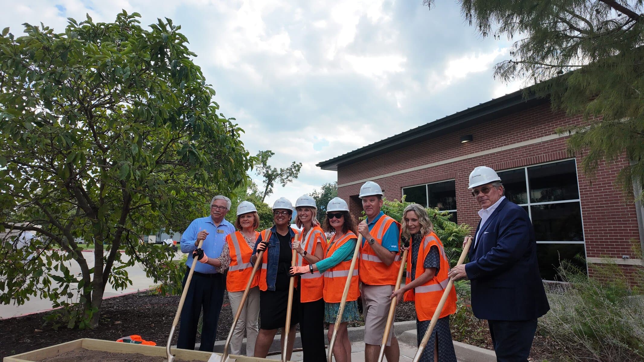 A group of people wearing hard hats and orange safety vests stand holding shovels at a groundbreaking ceremony in front of a brick building.